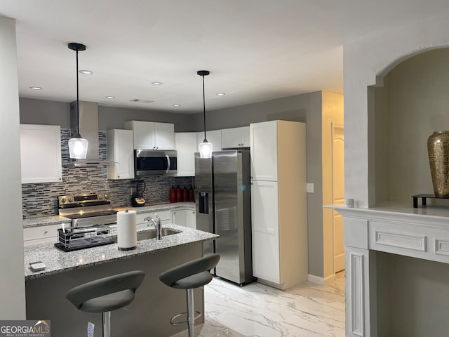 kitchen with light stone counters, hanging light fixtures, wall chimney range hood, white cabinetry, and appliances with stainless steel finishes