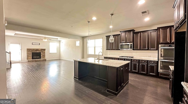 kitchen featuring dark hardwood / wood-style flooring, a fireplace, a center island with sink, appliances with stainless steel finishes, and ceiling fan