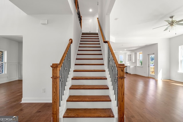 stairway featuring ceiling fan, a decorative wall, wood finished floors, baseboards, and wainscoting