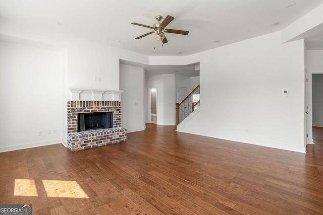 unfurnished living room with baseboards, dark wood finished floors, ceiling fan, stairway, and a brick fireplace