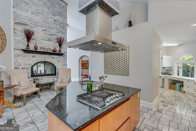 kitchen with a stone fireplace, dark stone countertops, a towering ceiling, light tile patterned floors, and island exhaust hood