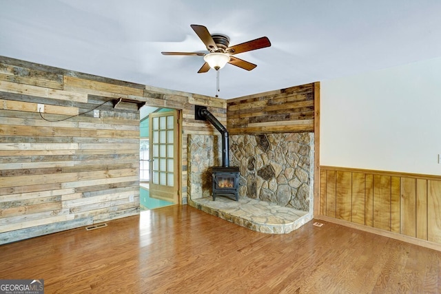 unfurnished living room featuring a wood stove, ceiling fan, hardwood / wood-style floors, and wooden walls