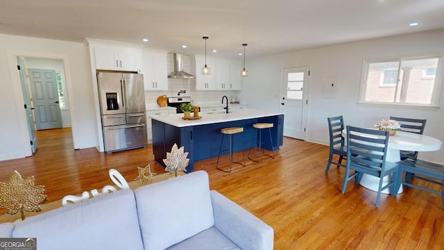 kitchen featuring light wood-type flooring, an island with sink, decorative backsplash, stainless steel fridge with ice dispenser, and wall chimney exhaust hood