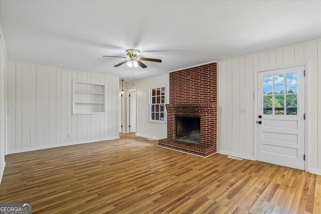 unfurnished living room featuring wood-type flooring, a brick fireplace, wooden walls, ceiling fan, and built in features