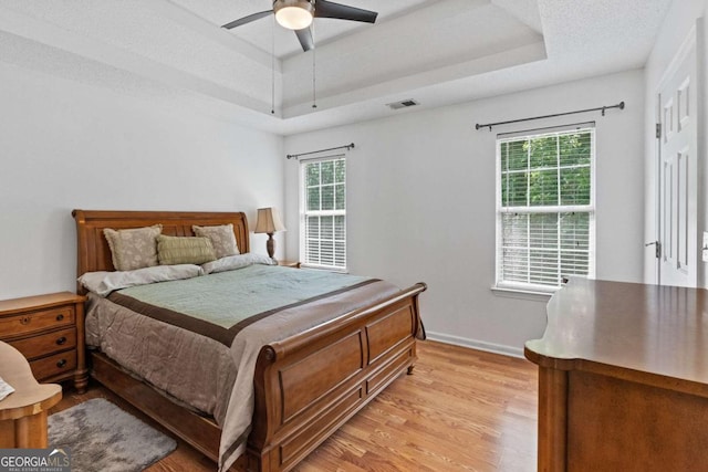 bedroom with a textured ceiling, ceiling fan, light wood-type flooring, and a tray ceiling