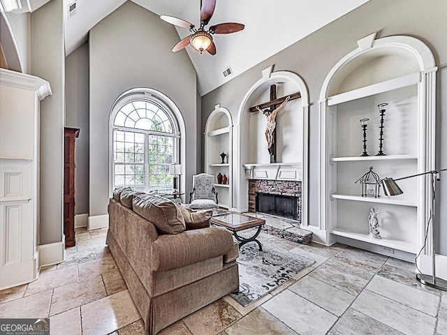 living room with built in shelves, high vaulted ceiling, a fireplace, a ceiling fan, and stone tile floors