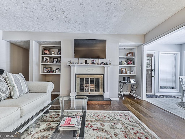 living room featuring built in features, a glass covered fireplace, a textured ceiling, wood finished floors, and baseboards