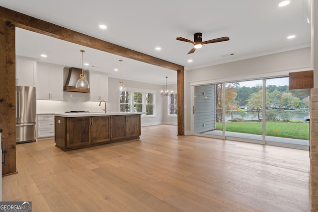 kitchen with light wood-type flooring, wall chimney exhaust hood, decorative light fixtures, a center island with sink, and stainless steel refrigerator