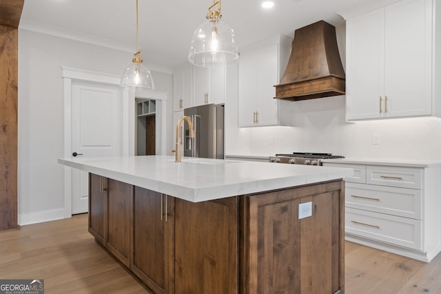 kitchen with custom exhaust hood, stainless steel appliances, a center island with sink, light hardwood / wood-style floors, and white cabinetry