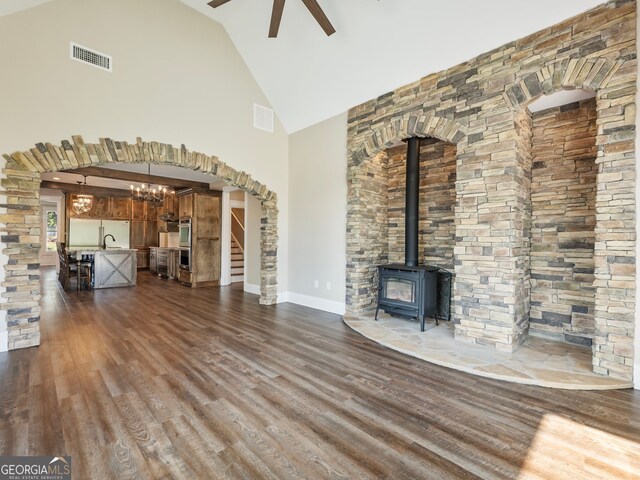 unfurnished living room with dark hardwood / wood-style floors, ceiling fan with notable chandelier, a wood stove, sink, and high vaulted ceiling