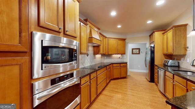 kitchen featuring premium range hood, light hardwood / wood-style floors, stainless steel appliances, sink, and dark stone counters