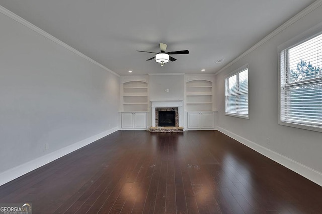 unfurnished living room with built in shelves, ceiling fan, a stone fireplace, dark hardwood / wood-style flooring, and crown molding