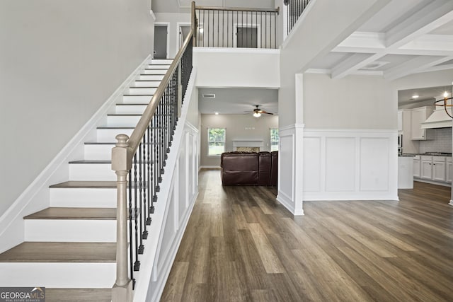 stairway with wood finished floors, coffered ceiling, beam ceiling, a towering ceiling, and wainscoting