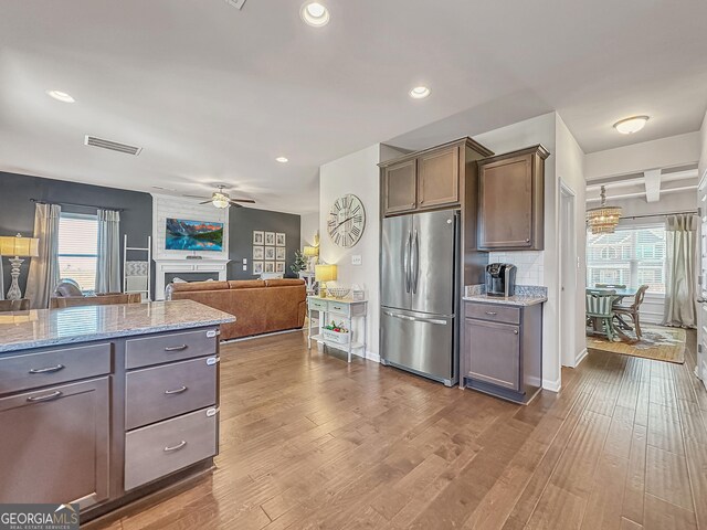 kitchen with a fireplace, light wood-type flooring, ceiling fan with notable chandelier, stainless steel fridge, and light stone counters