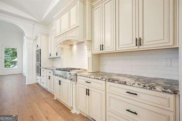 kitchen featuring appliances with stainless steel finishes, ornate columns, cream cabinetry, light wood-type flooring, and dark stone counters