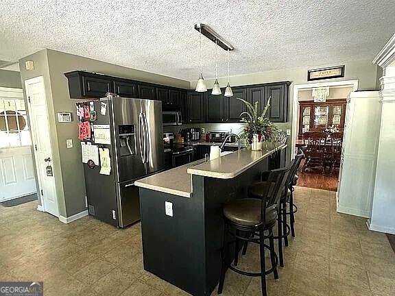 kitchen featuring a textured ceiling, a kitchen breakfast bar, appliances with stainless steel finishes, an island with sink, and hanging light fixtures