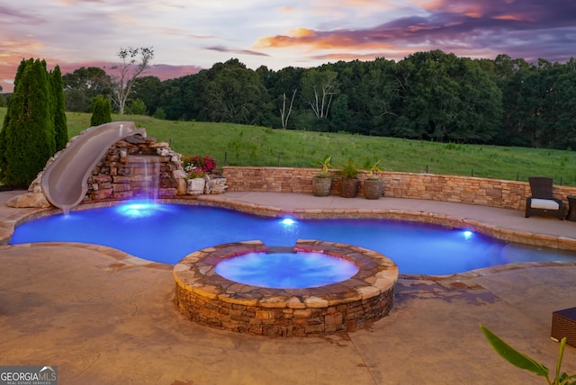 pool at dusk featuring a water slide, pool water feature, an in ground hot tub, and a patio area