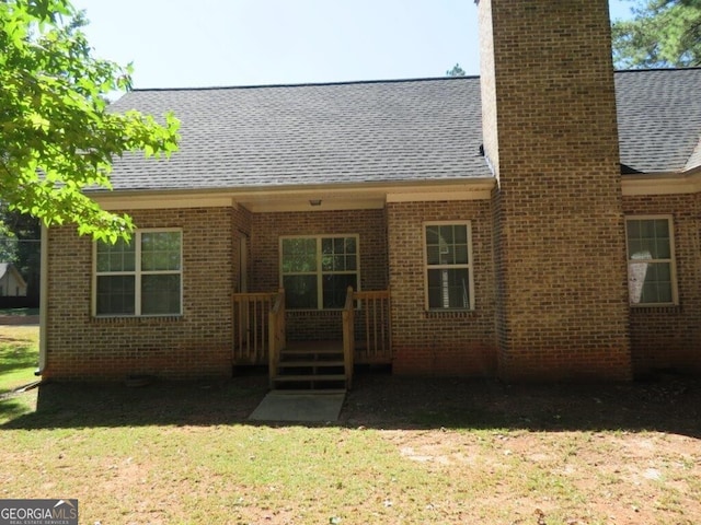 rear view of house featuring a lawn, brick siding, roof with shingles, and a chimney