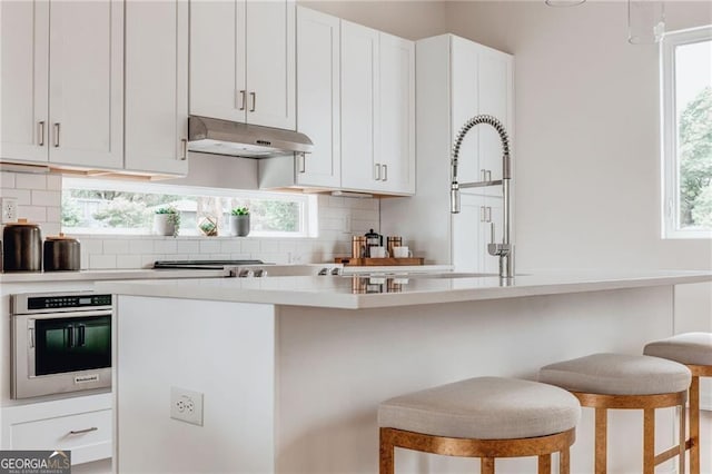 kitchen featuring a wealth of natural light, oven, under cabinet range hood, backsplash, and a breakfast bar area