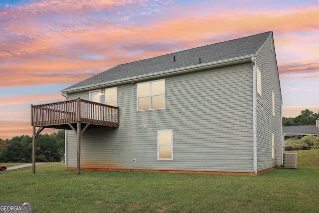 back of house at dusk featuring a wooden deck, a lawn, and roof with shingles