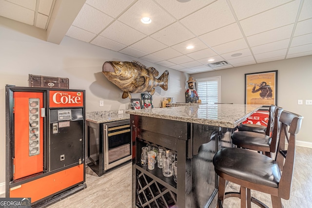 bar featuring beverage cooler, a paneled ceiling, light stone countertops, and light hardwood / wood-style flooring