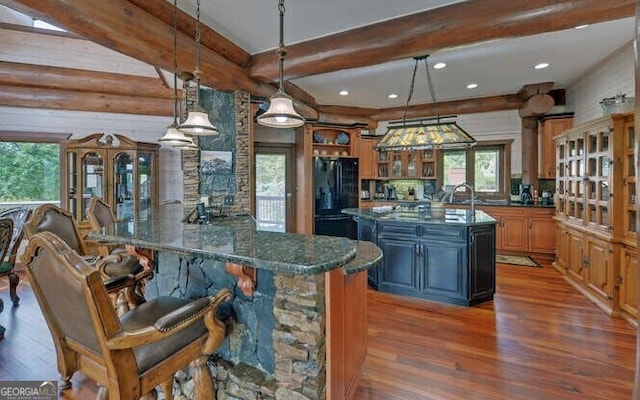 kitchen featuring black refrigerator, log walls, beam ceiling, hanging light fixtures, and an island with sink