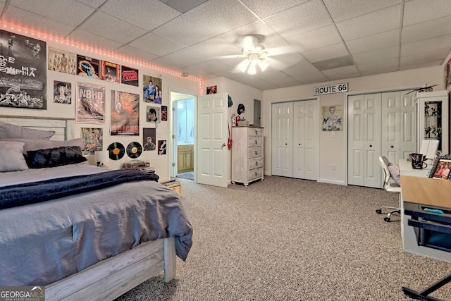 bedroom featuring ceiling fan, ensuite bathroom, a paneled ceiling, carpet, and two closets