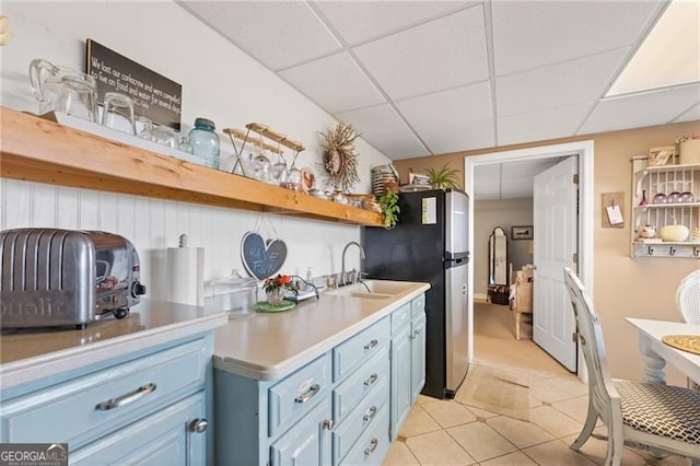 kitchen featuring stainless steel fridge, blue cabinetry, light tile patterned floors, sink, and a drop ceiling