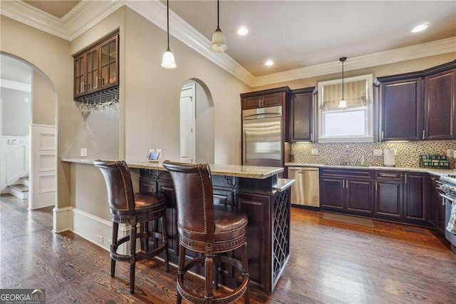 kitchen with pendant lighting, dark wood-type flooring, stainless steel appliances, ornamental molding, and light stone counters