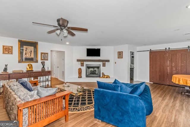 living area featuring wood finished floors, ceiling fan, a barn door, a fireplace with raised hearth, and recessed lighting