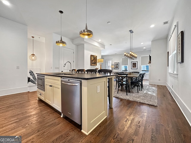 kitchen with a kitchen island with sink, stainless steel appliances, sink, hanging light fixtures, and dark wood-type flooring