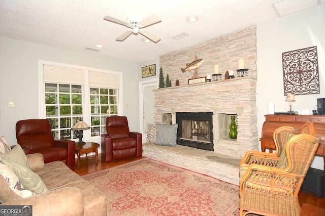 living room featuring ceiling fan, a stone fireplace, hardwood / wood-style flooring, and a textured ceiling