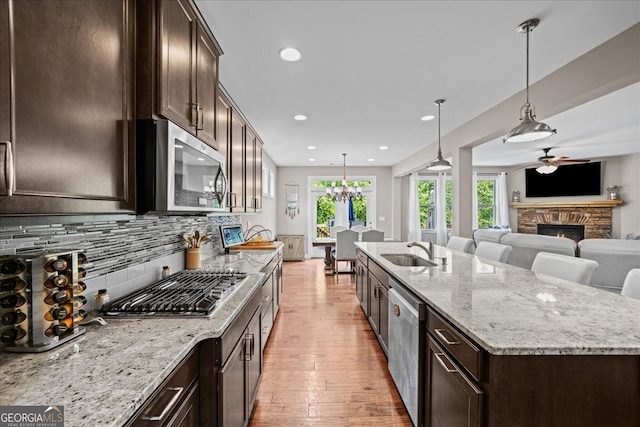 kitchen featuring a stone fireplace, an island with sink, hanging light fixtures, stainless steel appliances, and light stone counters