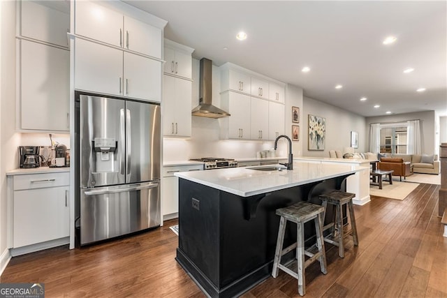 kitchen with an island with sink, sink, wall chimney exhaust hood, appliances with stainless steel finishes, and dark hardwood / wood-style flooring