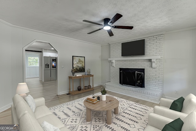 living room with ceiling fan, a brick fireplace, light hardwood / wood-style floors, a textured ceiling, and ornamental molding