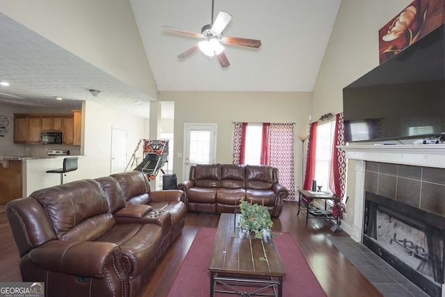 living room featuring dark wood-type flooring, ceiling fan, a tiled fireplace, and high vaulted ceiling