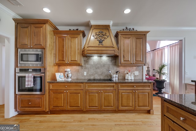 kitchen with dark stone counters, ornamental molding, stainless steel appliances, custom range hood, and light hardwood / wood-style floors