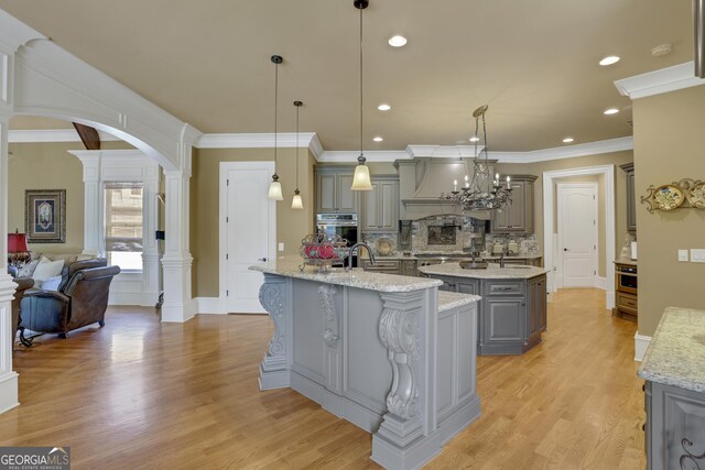 kitchen with light wood-type flooring, stainless steel double oven, hanging light fixtures, ornate columns, and a center island with sink