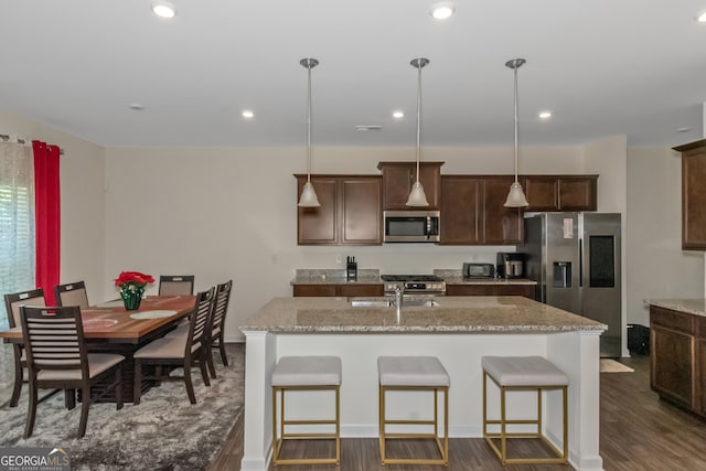 kitchen with decorative light fixtures, light stone counters, stainless steel appliances, dark hardwood / wood-style flooring, and a kitchen island with sink