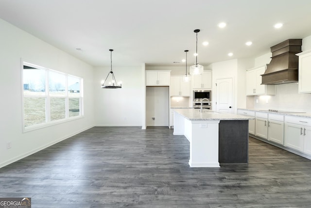 kitchen with premium range hood, white cabinetry, decorative light fixtures, a center island with sink, and stainless steel microwave