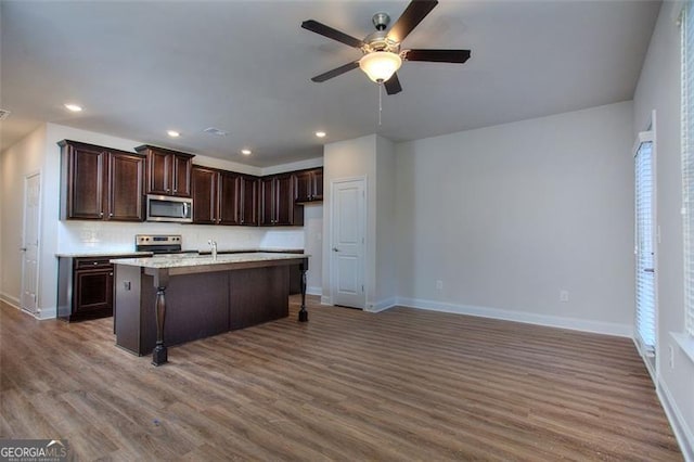 kitchen with dark brown cabinets, an island with sink, appliances with stainless steel finishes, hardwood / wood-style floors, and ceiling fan