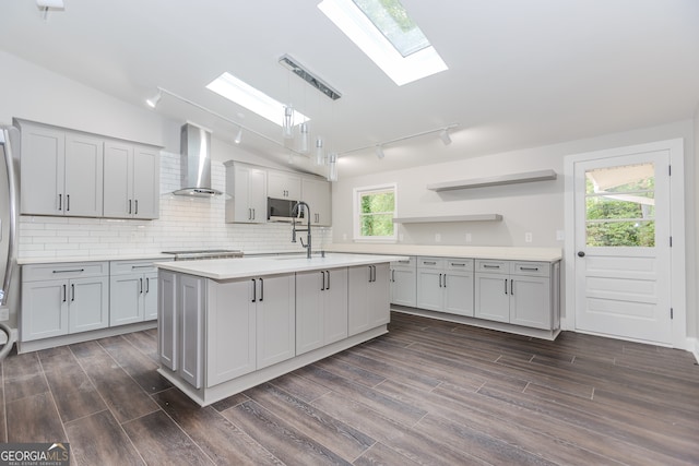 kitchen featuring gray cabinetry, vaulted ceiling with skylight, dark hardwood / wood-style floors, wall chimney exhaust hood, and a center island with sink