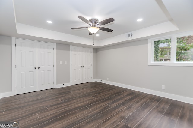 unfurnished bedroom featuring dark wood-type flooring, ceiling fan, a raised ceiling, and multiple closets