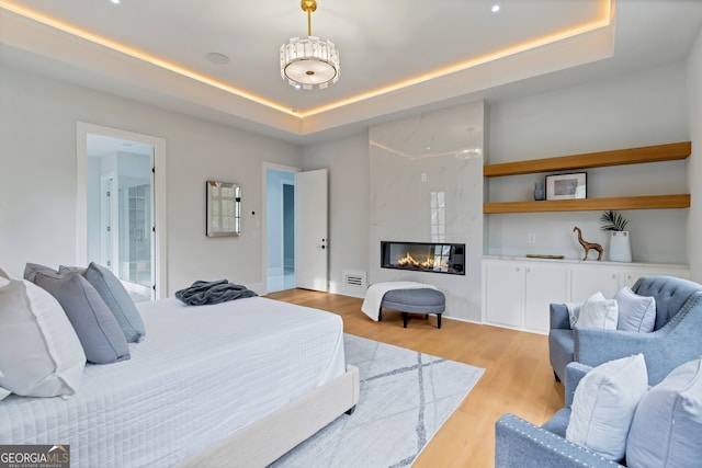 bedroom featuring light wood-type flooring, a tray ceiling, connected bathroom, and a premium fireplace