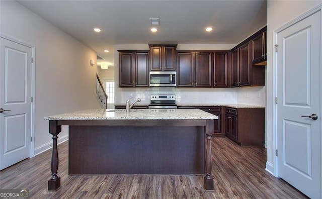 kitchen with appliances with stainless steel finishes, decorative backsplash, dark brown cabinets, an island with sink, and light stone countertops