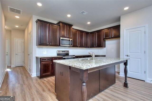 kitchen with light stone counters, light hardwood / wood-style floors, sink, a center island with sink, and appliances with stainless steel finishes