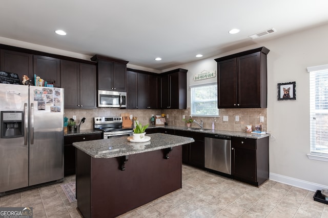 kitchen featuring light stone countertops, tasteful backsplash, a center island, stainless steel appliances, and dark brown cabinetry