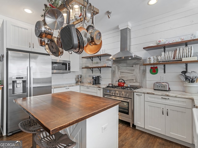 kitchen featuring dark wood-type flooring, light stone countertops, appliances with stainless steel finishes, white cabinets, and wall chimney range hood