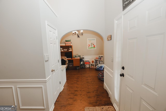 hallway featuring a decorative wall, arched walkways, dark wood-style floors, an inviting chandelier, and wainscoting