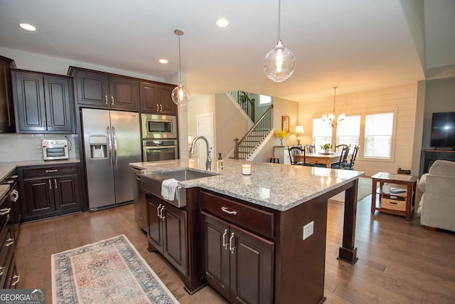 kitchen with decorative light fixtures, dark wood-type flooring, appliances with stainless steel finishes, a center island with sink, and dark brown cabinetry